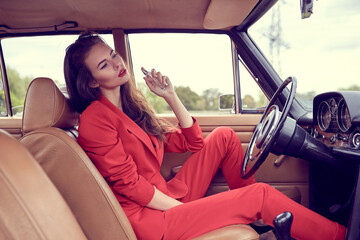 Beautiful young vogue woman in red suit sitting in retro car using a perfume.
