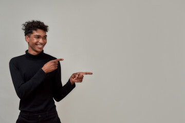 Portrait of cheerful young guy dressed elegantly looking happy while pointing at right side, presenting something, standing isolated over gray background