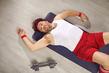 Funny tired fat man lying on fitness mat after gym workout. Exhausted guy in retro sweatband, tank...