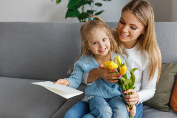 International women's and mother's day. Little cute daughter with bouquet of yellow flowers and postcard congratulates her smiling mom. Happy family holidays at home. Curly girl hugging mommy.