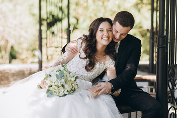 Stylish groom in a gray checkered suit and a cute blonde bride in a white lace dress with a long veil are hugging in the park near the old house. Wedding portrait of smiling newlyweds.