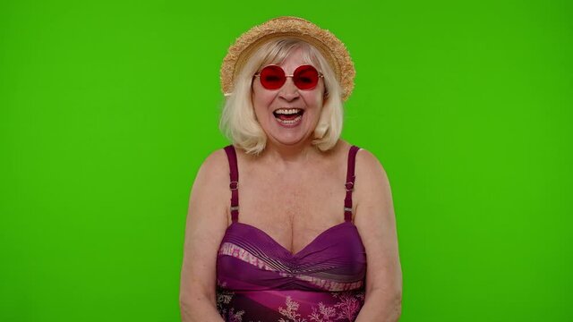 Senior woman traveler in red sunglasses, looking at camera, smiling, starting laughing. Mature old grandmother in swimsuit bra on chroma key background. Tourism, summer holiday vacation, trip to sea