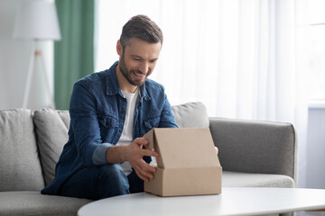 Happy man unpacking delivery box, home interior - obrazy, fototapety, plakaty