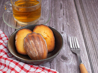 Madeleine homemade traditional French small cookies on black a dish with a cloth and a teacup placed on a wooden background. Lemon glaze madeleines. Seashell sweet cakes