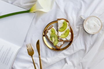 Breakfast in bed on white sheets with flower and book, a cup o coffee and sweet tost. Concept spring top view. Copy space