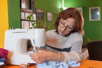 An adult woman sitting at home in the living room during the day on her sewing machine during day time, wearing glasses and a sweater