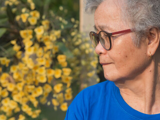 Senior woman with short gray hair wearing glasses, smiling and looking at yellow orchids while standing in a garden. Side view. Space for text. Concept of aged people and healthcare