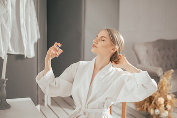 Young blonde beautiful woman in the white bathrobe sitting next to the table and using perfume bottle