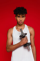 sportive african american man in tank top working out with dumbbell isolated on red