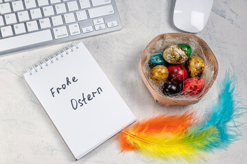 The words in German Frohe Ostern or Happy Easter in a notebook on the desktop with colored eggs in a nest and colored feathers. Easter in the office.