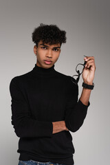 young african american man in turtleneck sweater holding glasses while posing isolated on grey