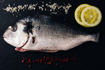 Fresh uncooked dorado or sea bream ready to cook. Raw fish with ingredients and seasoning like  rosemary , lemon, salt, black pepper. black background. Cooking food for healthy lunch or dinner