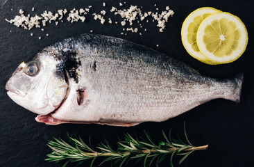 Fresh uncooked dorado or sea bream ready to cook. Raw fish with ingredients and seasoning like  rosmary , lemon, salt. black background. Cooking food for healthy lunch or dinner. top view