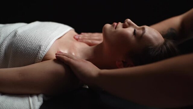 Close-up face of young relaxed woman lying down on massage table with close eyes during shoulder and neck massage at spa salon. Male masseur professionally massaging shoulders on black background.