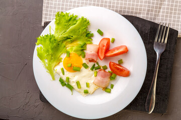 Scrambled eggs with bacon garnished with tomato and green onions and salad on a gray concrete background on a wooden stand next to a fork.