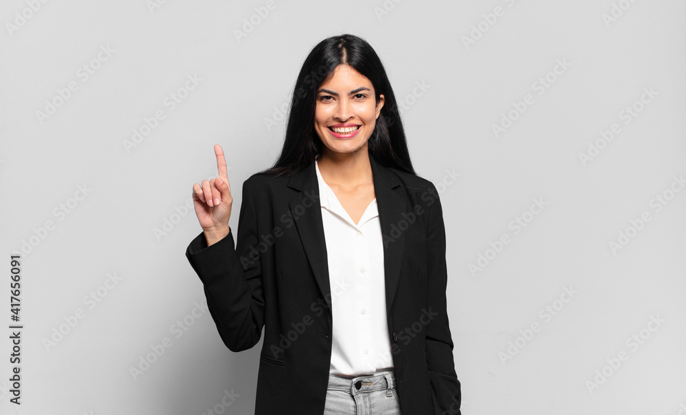 Wall mural young hispanic businesswoman smiling cheerfully and happily, pointing upwards with one hand to copy space