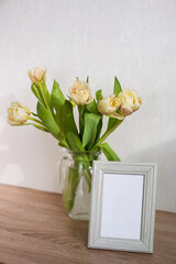 Bouquet of beautiful tulips and empty frame mock up. Copy space. International Women's Day celebration. White wall background. Scandinavian interior.