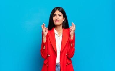 young hispanic businesswoman crossing fingers anxiously and hoping for good luck with a worried look