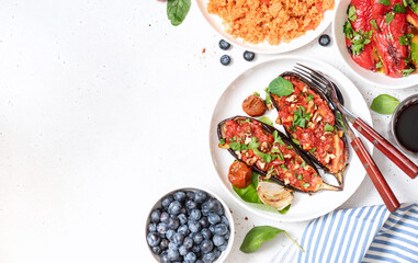 Various cooked vegan food in plates on a white concrete table top view. Eggplant with tomato sauce, couscous, peppers, berries, spinach salad. Vegetarian lunch or dinner. Copy space for text