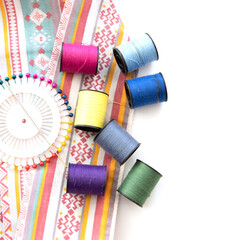 Selective Focus of colored needles and thread reel on patterned fabric placed on the left side, copy space, white background, top view, sewing and embroidery concept. 