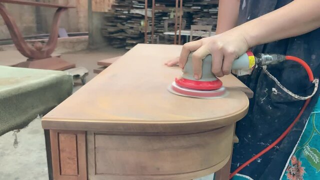 A mechanic woman using a red pneumatic round orbital sander. Work on a mahogany table The dust produced by this process floats in the air.