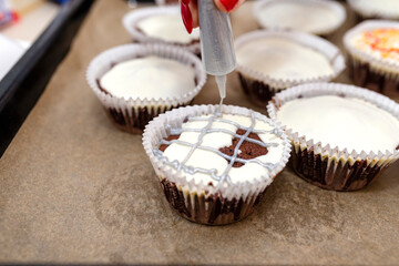 Chocolate-brown cupcakes wrapped in white paper and covered with white frosting with colorful decorations, visible fingers and nails.