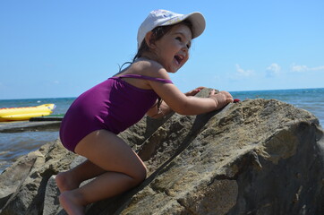 Little cute girl in a purple swimsuit crawls on a huge stone by the sea. The concept of vacation, childhood, vacation.