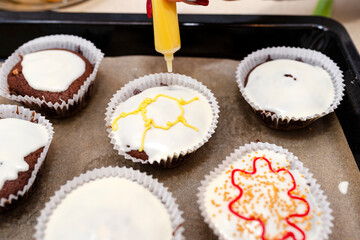 A woman squeezes colored frosting from a tube onto chocolate brown cupcakes covered with white frosting with colorful decorations.