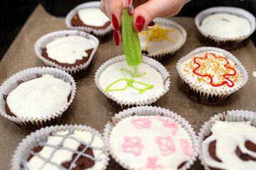 A woman squeezes colored frosting from a tube onto chocolate brown cupcakes covered with white frosting with colorful decorations.