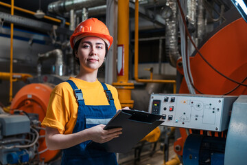 Portrait of female smiling worker in uniform and helmet with a folder and a pen in her hand. In the background-boiler room