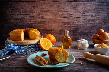 Portion of homemade orange sponge cake in a light turquoise plate accompanied by the ingredients for its preparation on a rustic wooden table, traditional pastry concept. Wooden background.