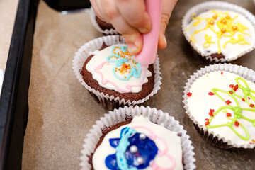 A child squeezes colored frosting from a tube onto chocolate brown cupcakes covered with white frosting with colorful decorations.