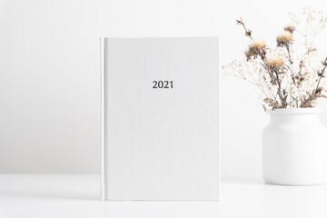 White book mockup, diary for 2021, white table, dried flowers in vase. Front view. Place for text,...