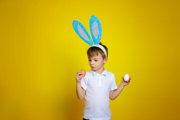 A boy dressed in a white shirt with short sleeves and bunny ears chooses which egg to paint for Easter. The child stands on a yellow background.