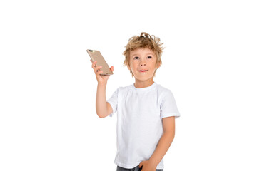 A little boy holds a phone in his hand and looks up happily with a smile. Boy stands on a white background, dressed in a white t-shirt