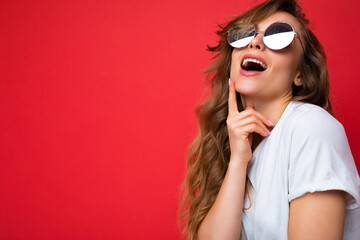 Shot of beautiful thinking young dark blonde curly woman isolated over red background wall wearing casual white t-shirt and stylish sunglasses looking up and having an idea