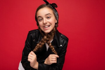 Photo shot of pretty positive smiling brunette little female teenager with pigtails wearing stylish black leather jacket and white t-shirt standing isolated over red background wall looking at camera