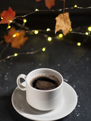 Close-up of a white cup of aromatic espresso coffee on a dark background with hanging dry maple leaves and a garland