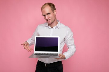 Photo shot of handsome smiling blonde man holding computer laptop with empty monitor screen with mock up and copy space wearing white shirt looking at camera pointing at netbook isolated over pink