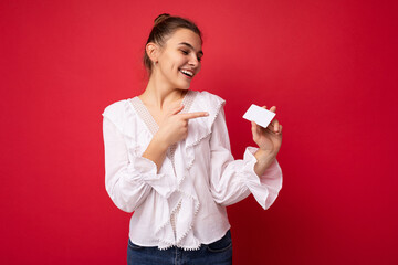 Photo shot of beautiful positive smiling young dark blonde female person wearing white blouse isolated over red background holding credit card looking and pointing finger at plastic contactless card