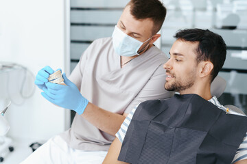 Male dentist in medical mask explaining artificial teeth to smiling patient in clinic