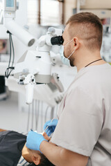 Young man with patient bib on a dental chair and a dentist who sits next to him. He looks on his teeth using a dental microscope and holds a dental bur and a mirror.