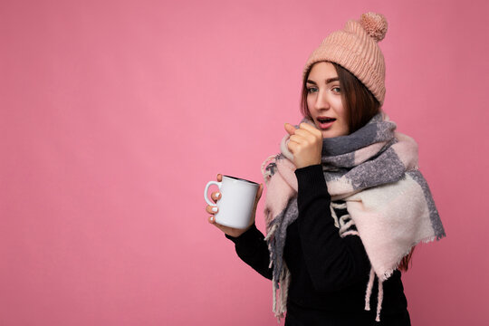 Beautiful sick young brunet woman wearing black sweater hat and warm scarf isolated over pink background holding paper mug for mockup drinking hot tea and looking at camera and caughing