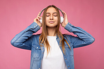 Beautiful happy with sincere emotions young blonde woman wearing blue jean shirt and white t-shirt isolated over pink background wearing white wireless bluetooth headphones listening to cool music and