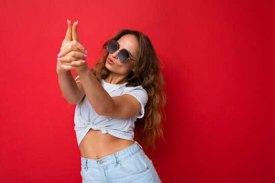 Photo shot of beautiful positive young blonde woman wearing casual clothes and stylish sunglasses isolated over colorful background looking to the side and showing gun gesture