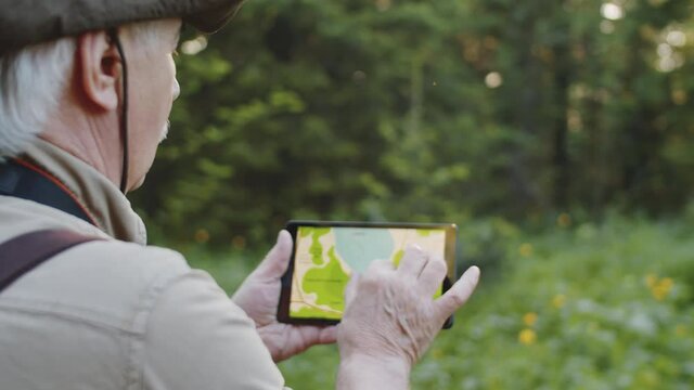Senior man standing in forest and examining map on digital tablet while hiking in wild nature
