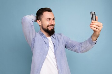 Handsome young brunet bearded man wearing casual stylish clothes standing isolated over blue background wall holding smartphone taking selfie photo looking at mobile phone screen display