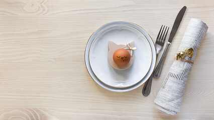 Easter breakfast on a wooden table: a boiled egg on a stand in the form of a cat, next to a napkin and cutlery: a fork and a knife