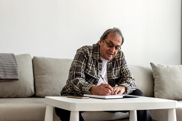 A senior Caucasian man in his 70s sitting at a table or desk, writing on a notepad at home. Senior grey hair man sitting in the living room and writing in his diary. Older man taking notes at home.