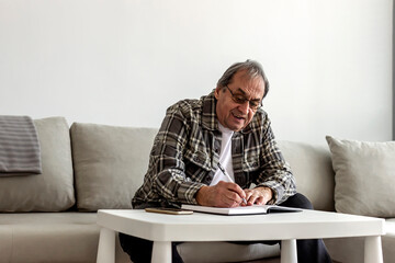 Obraz premium A senior Caucasian man in his 70s sitting at a table or desk, writing on a notepad at home. Senior grey hair man sitting in the living room and writing in his diary. Older man taking notes at home.